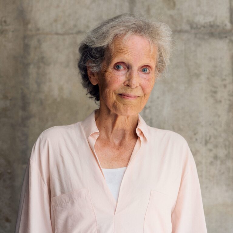 Ältere Frau mit grauen Haaren und blauen Augen, trägt ein helles Hemd, vor einer grauen Wand.