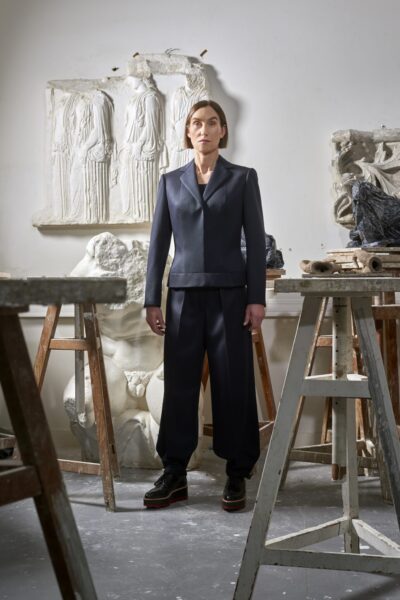 Mika'Ela Fisher steht in einem Atelier, umgeben von Skulpturen und Arbeitsbänken, in eleganter, dunkler Kleidung.