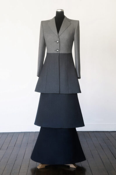 Graues, modisches Kleid mit taillierter Jacke und gestuften Rockteilen, auf einem schwarzen Kleiderständer.