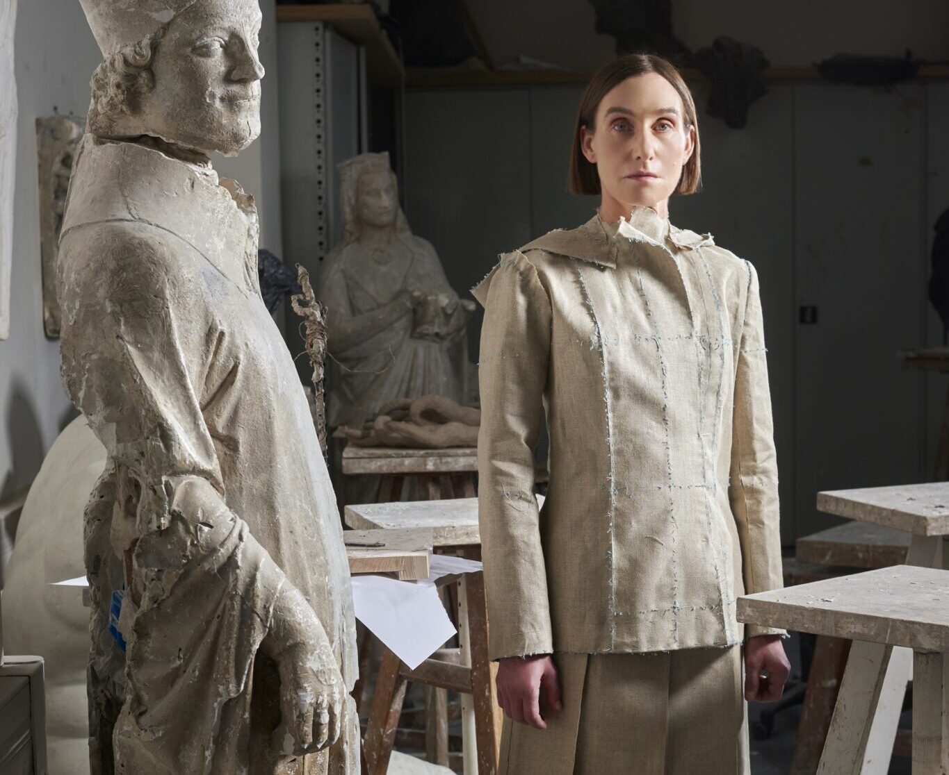 Frau in beiger Kleidung steht neben einer antiken Statue in einem Atelier mit Holzwerkbänken.