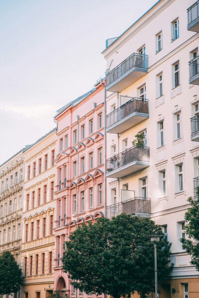 jonas-denil-HZPrWMk4wXY-unsplash_ - Relation Magazin Fassade von mehrstöckigen Wohngebäuden in sanften Farben mit Balkonen und Bäumen im Vordergrund.
